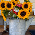 Bouquet of sunflowers and other flowers in a metal watering can on a wooden surface.