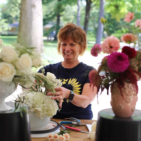 Woman arranging flowers at a table outdoors with trees in the background